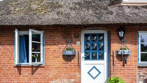 Una casa de ladrillo con techo de paja, una puerta blanca con detalles azules y dos cestas de flores colgantes.