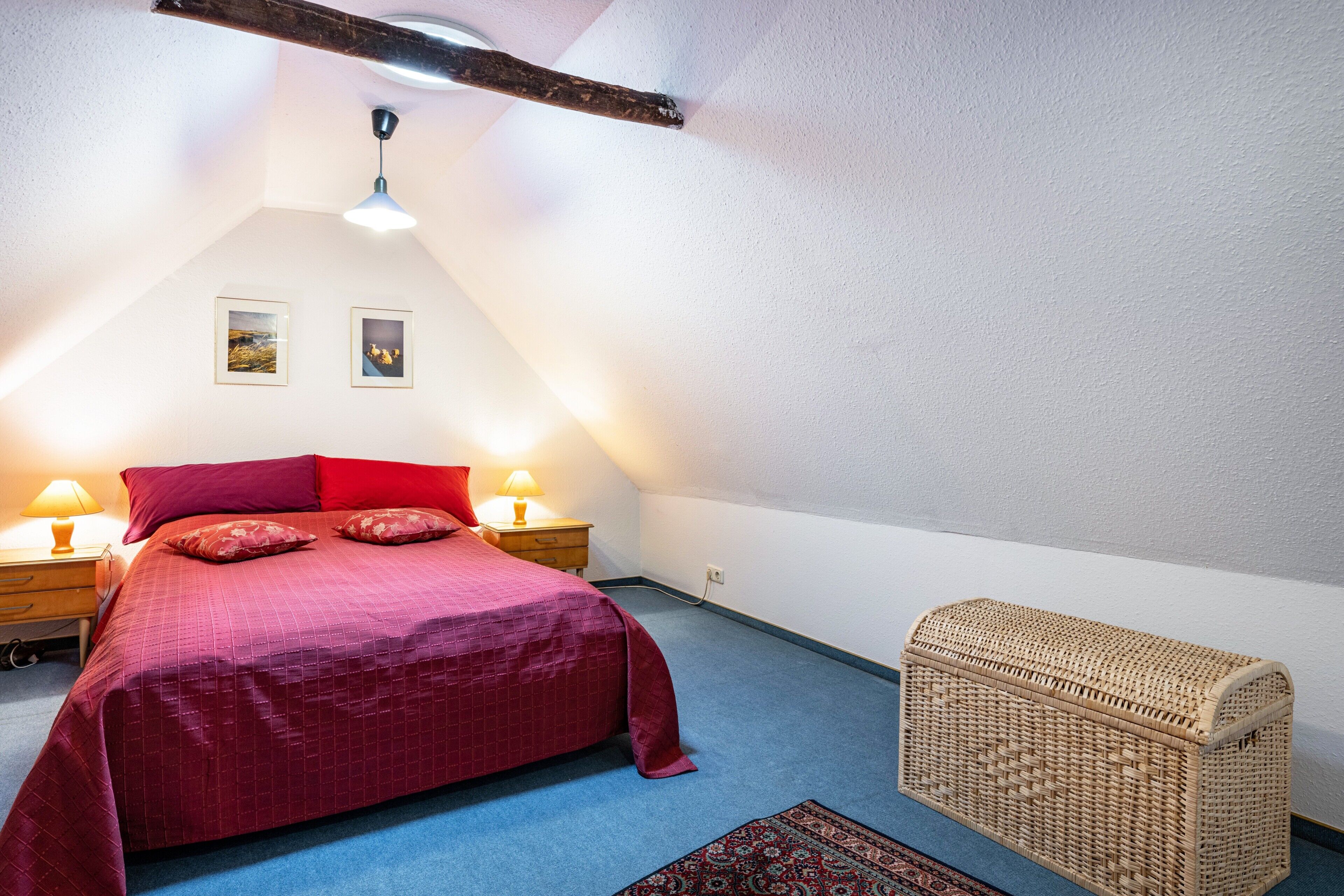 A bedroom with a bed, two bedside lamps, a red bedspread, a wicker chest, and a patterned rug.