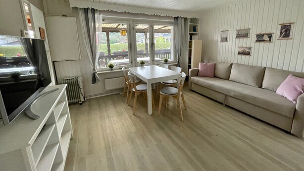 A room with a white ceiling, a dining table with chairs, a sofa, a television, and a window with curtains.