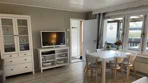A room with a dining table, chairs, a television on a stand, a cabinet with glass doors, and a window with curtains.