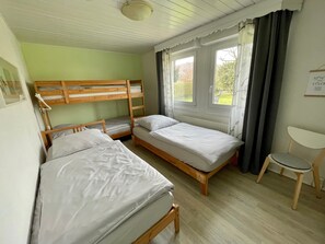 Una habitación con literas, una cama individual, una silla, un ventanal con cortinas y una lámpara en el techo.