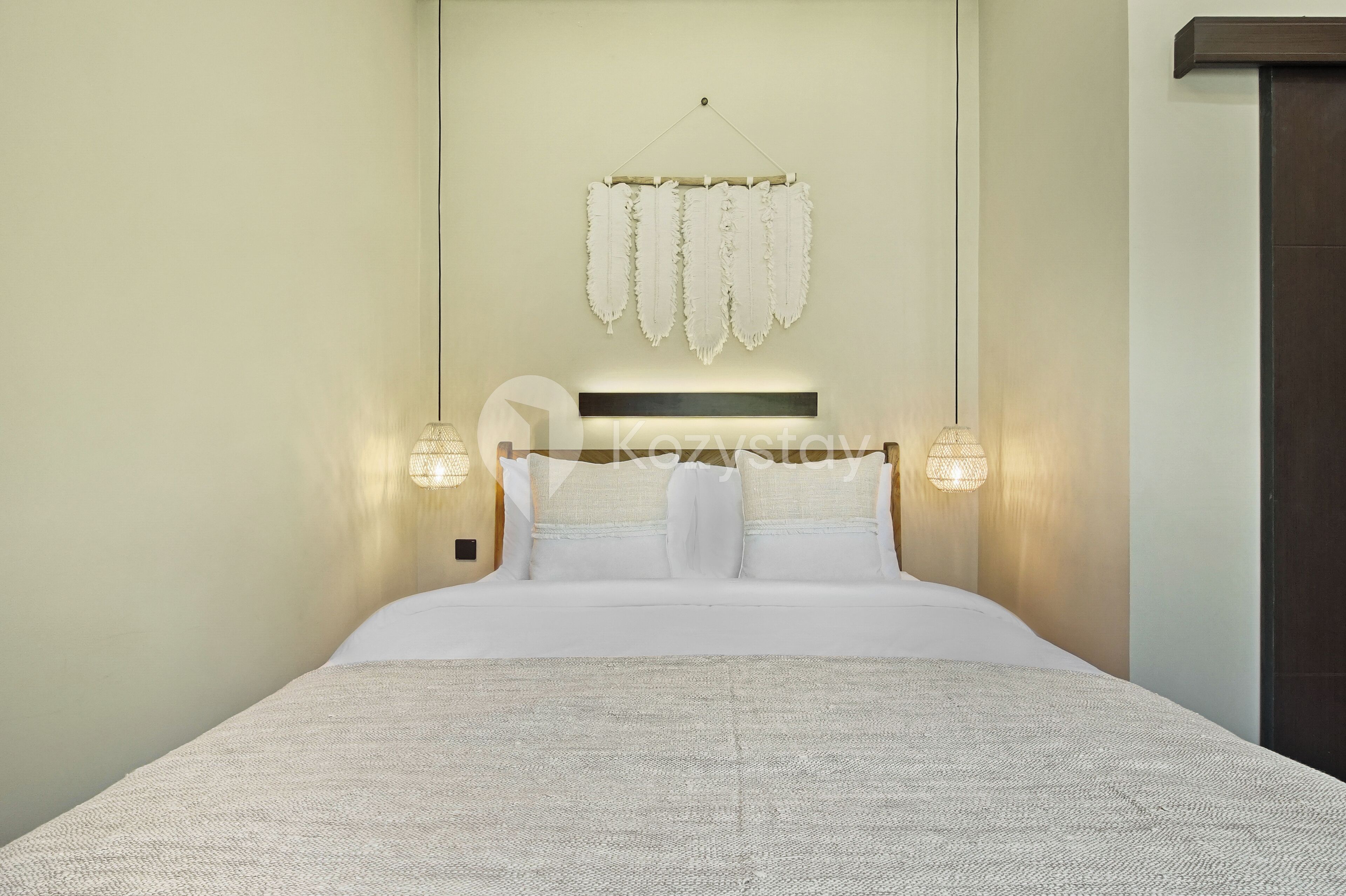 A neatly made bed with white linens and pillows, a hanging wall decoration, and two wall-mounted lamps.