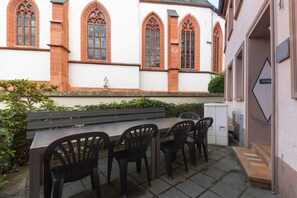 Un espace extérieur aménagé avec une table et des chaises, un banc, et un bâtiment doté de fenêtres en arc de cercle et d’une façade en briques.