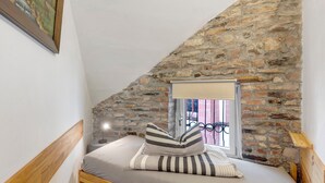 Un dormitorio con pared de piedra inclinada, una ventana con reja de metal, una cama con almohadas a rayas y un cabecero de madera.