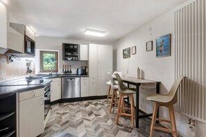 Une cuisine moderne avec des armoires blanches, un comptoir noir et un plancher à motifs en chevrons. On y trouve deux tabourets de bar et une table recouverte d’une nappe à motifs.