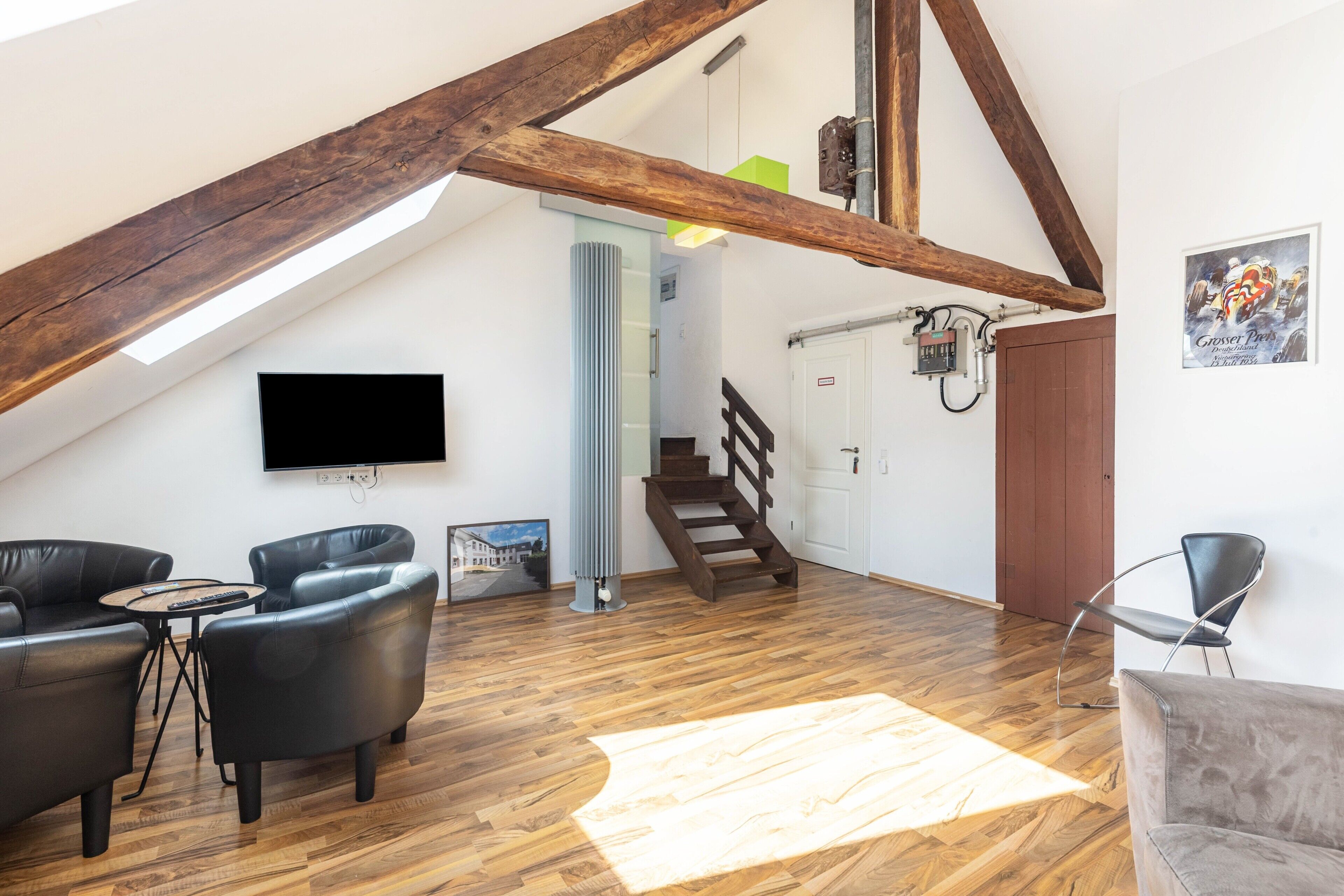 Habitación estilo loft con vigas de madera a la vista, un televisor montado en la pared, una escalera y un área de estar con sofás de cuero y una mesa de centro.