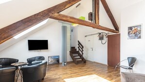 Een loft-achtige kamer met zichtbare houten balken, een aan de muur bevestigde televisie, een trap en een zithoek met leren banken en een salontafel.