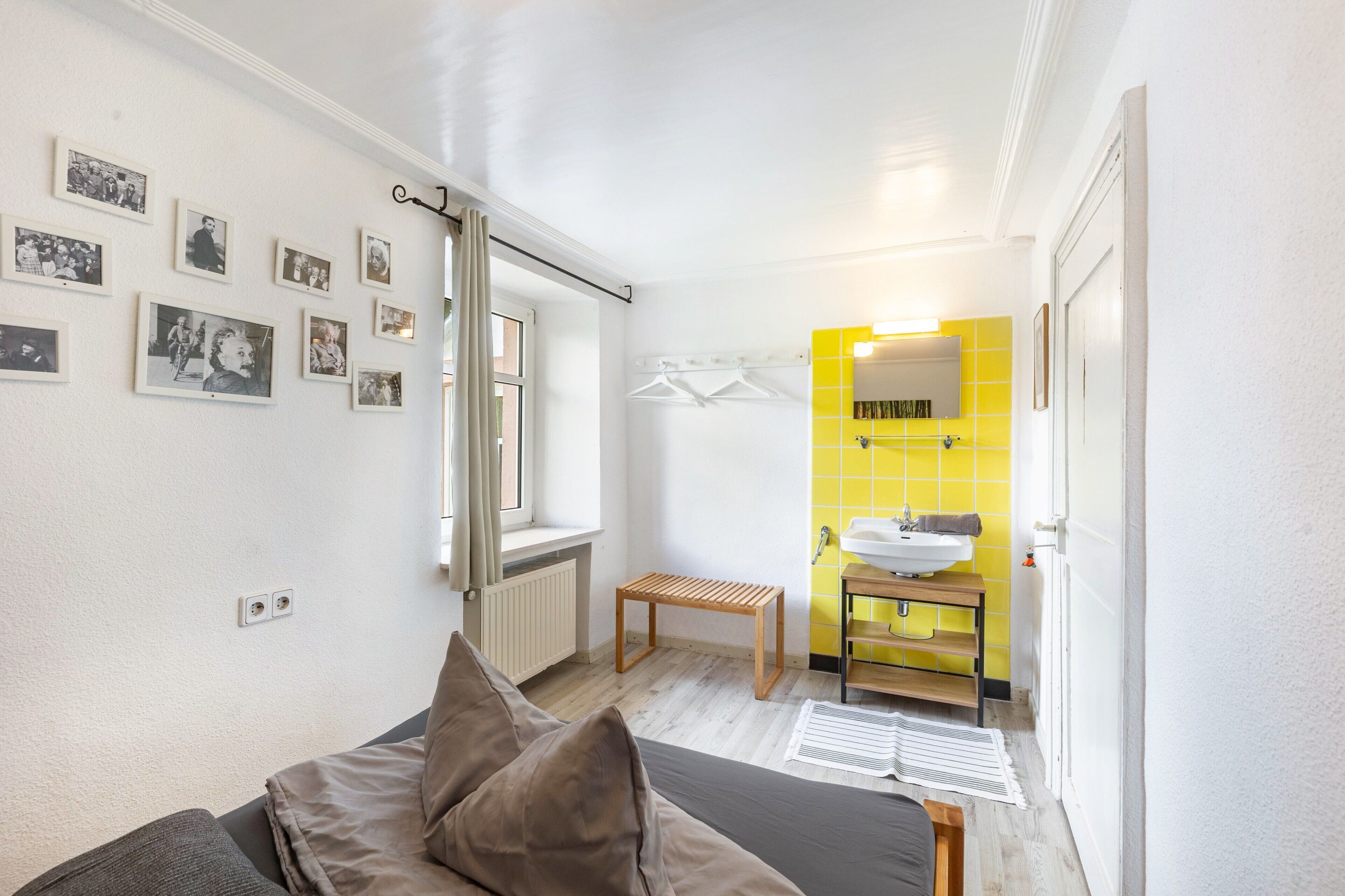 A bedroom with a bed, a bedside table, a window, and a bathroom with a sink and mirror.