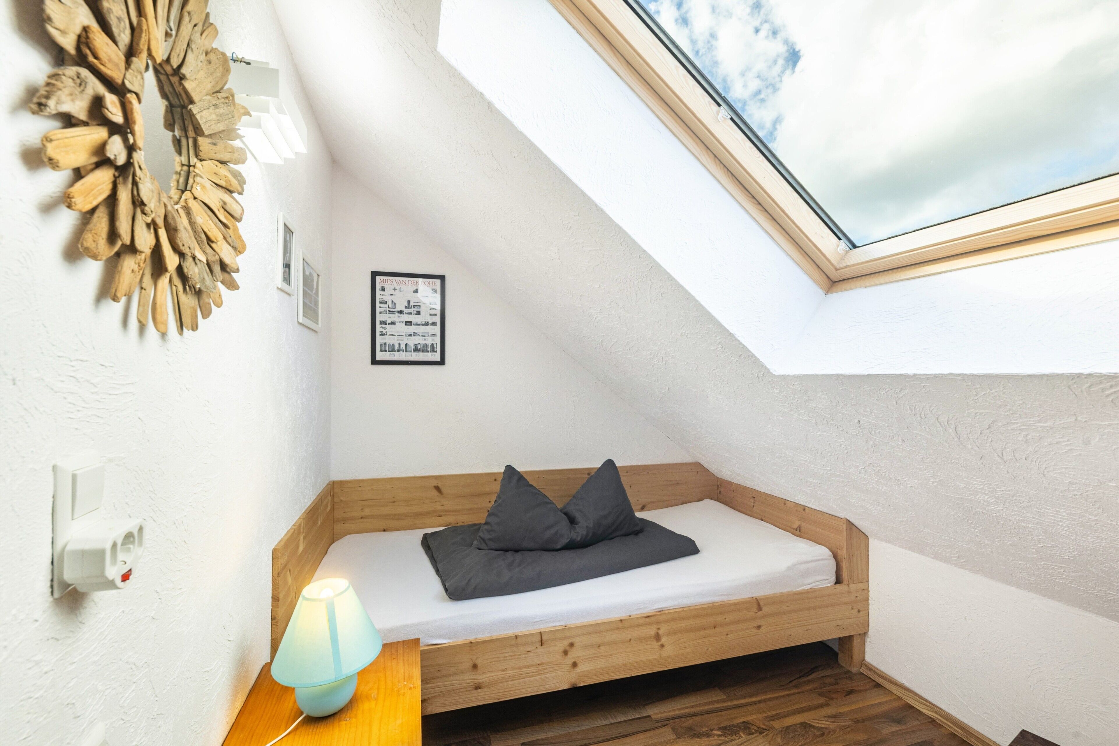Ein kleiner, gemütlicher Raum mit schrägem Dach, einem hölzernen Bett, einer Wandleuchte und einem gerahmten Bild.