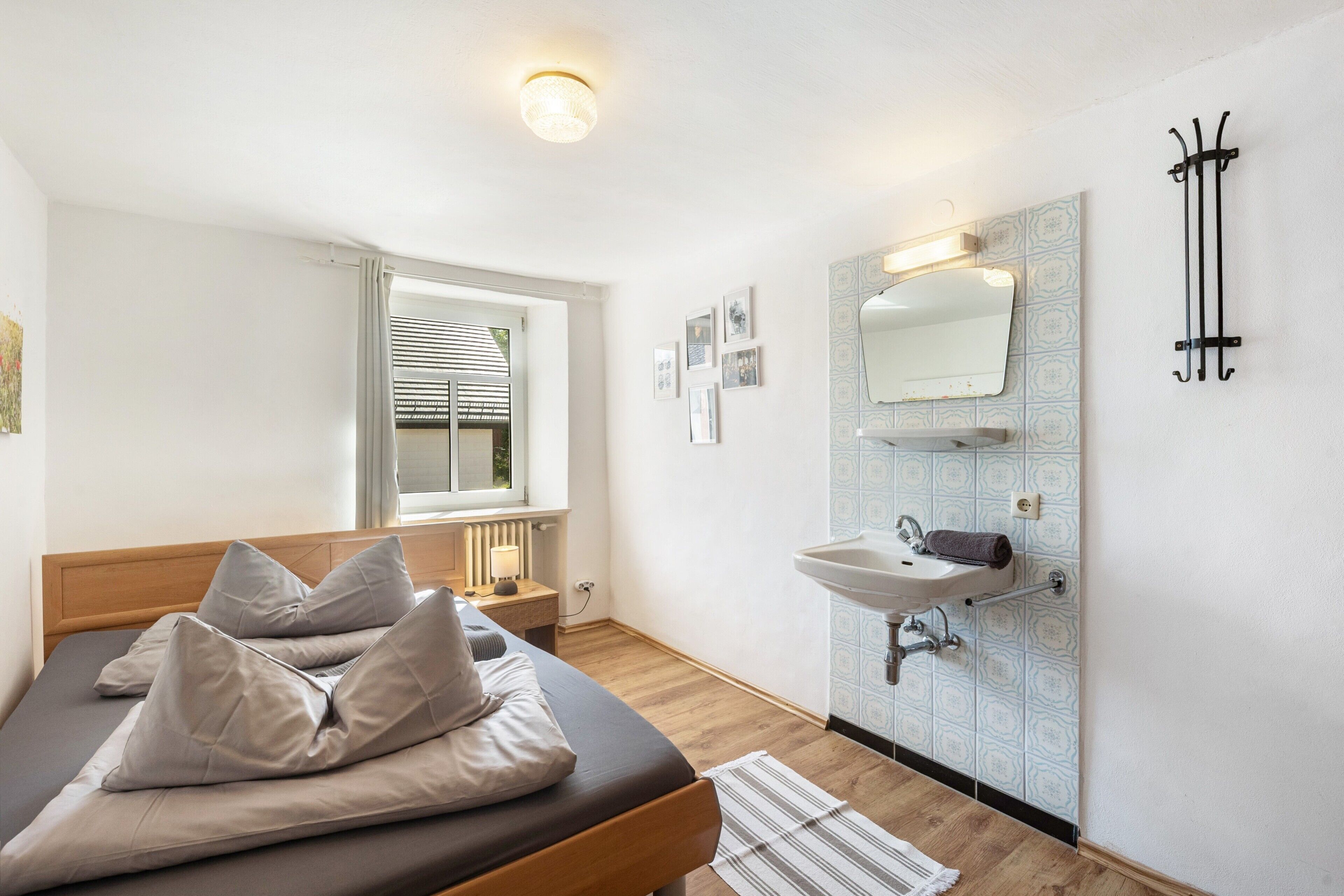 Un dormitorio con cama, mesita de noche, lámpara, ventana con persianas, y un baño con lavamanos y espejo.