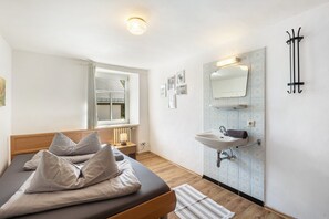 Ein Schlafzimmer mit Bett, Nachttisch, Lampe, Fenster mit Jalousien sowie ein Badezimmer mit Waschbecken und Spiegel.