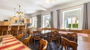 Een eethoek met een lange tafel, houten stoelen, een geruit tafelkleed en een keuken op de achtergrond.