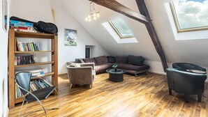 Een kamer in loftstijl met schuine plafonds, houten balken, een boekenkast, een bank, een bijzettafeltje en een stoel.