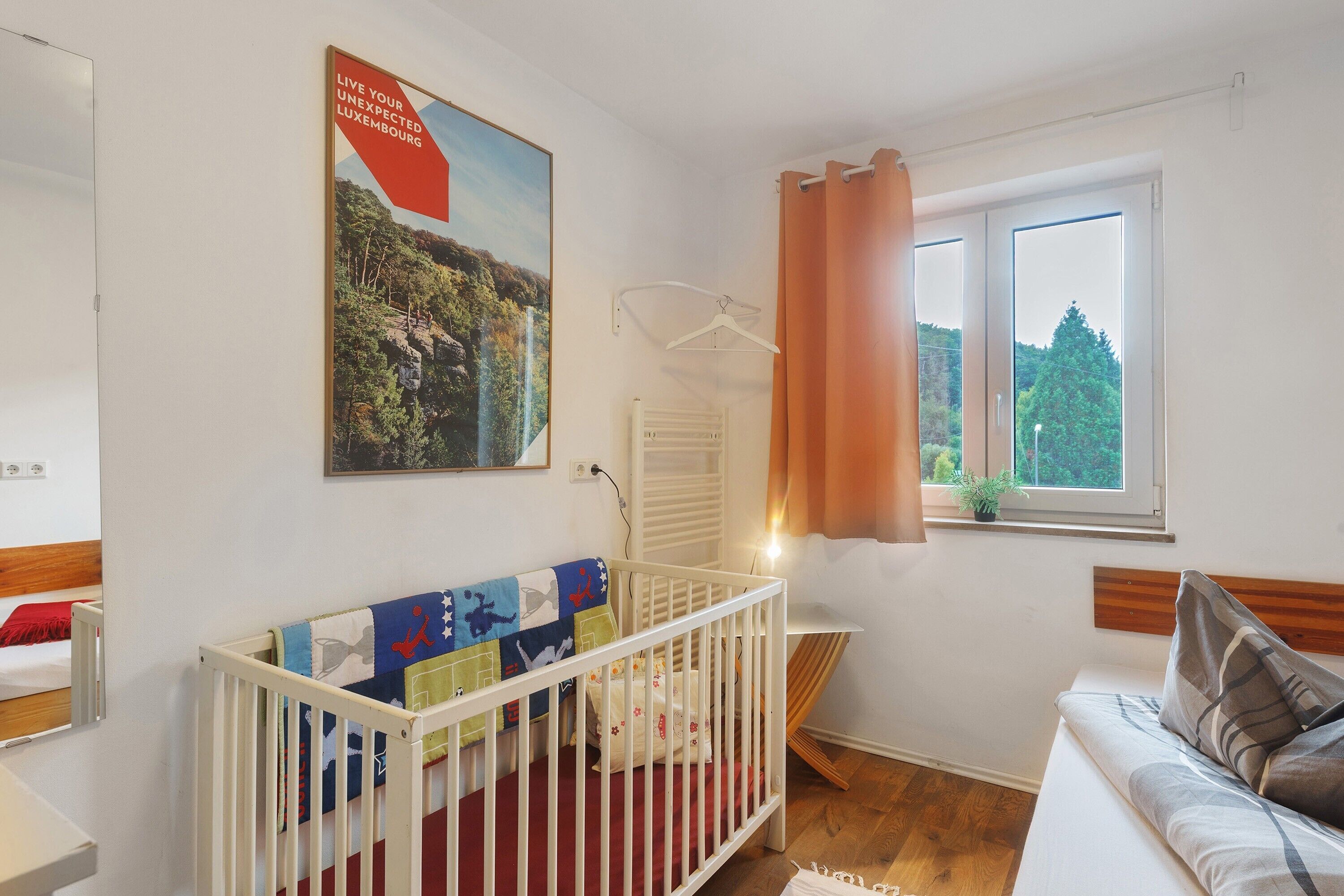 A room with a crib, a bed, a window with curtains, a radiator, and a poster on the wall.