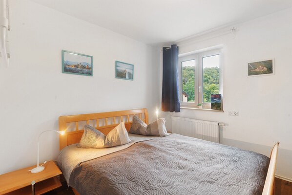 A bedroom with a wooden headboard, a bed with patterned bedding, a bedside table with a lamp, and a window with a view of greenery. - Holiday Home 'Eifel Atrium' with Private Terrace and Wi-Fi (Zweifelscheid)