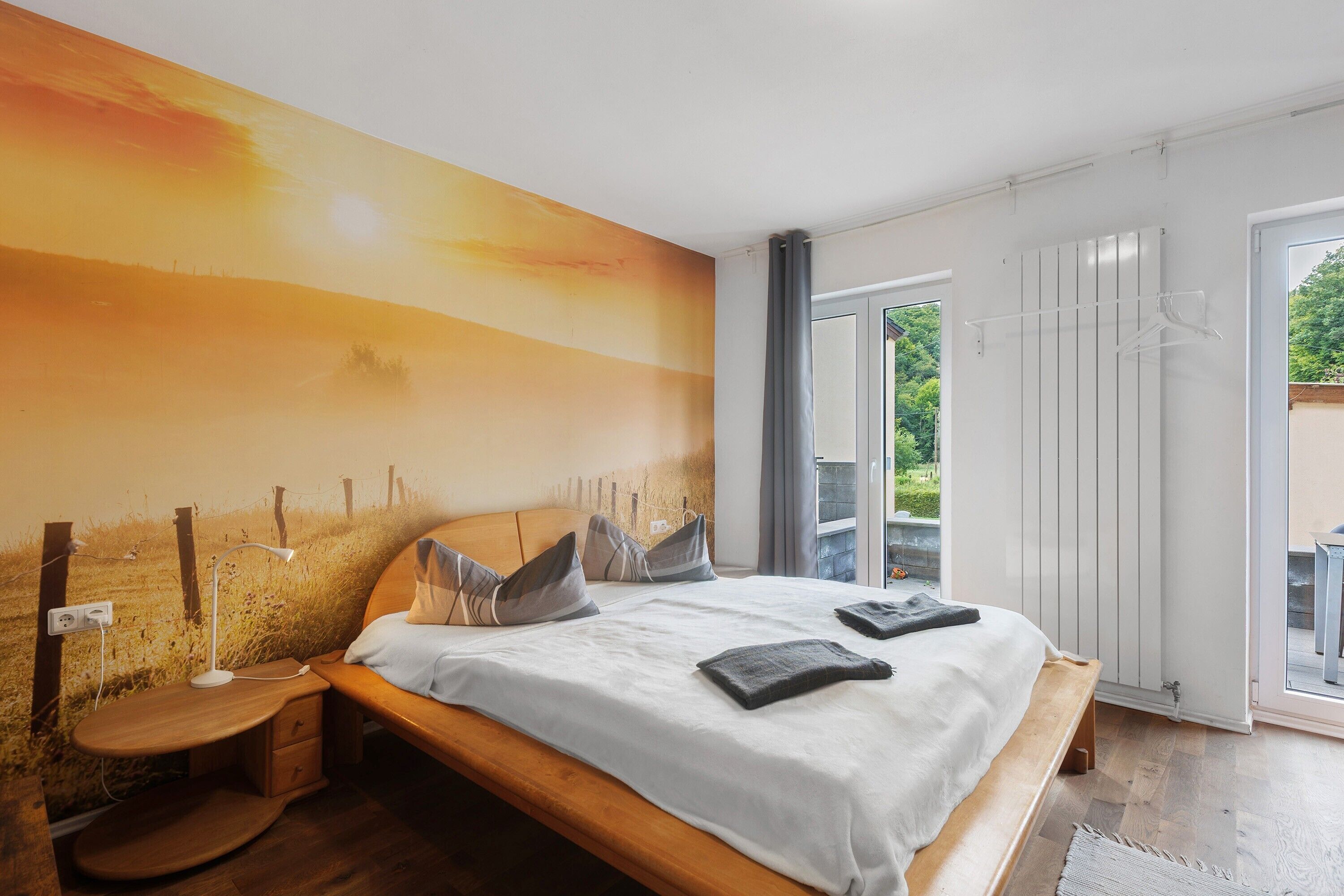 A bedroom with a bed, bedside table, and a view of greenery through a window.