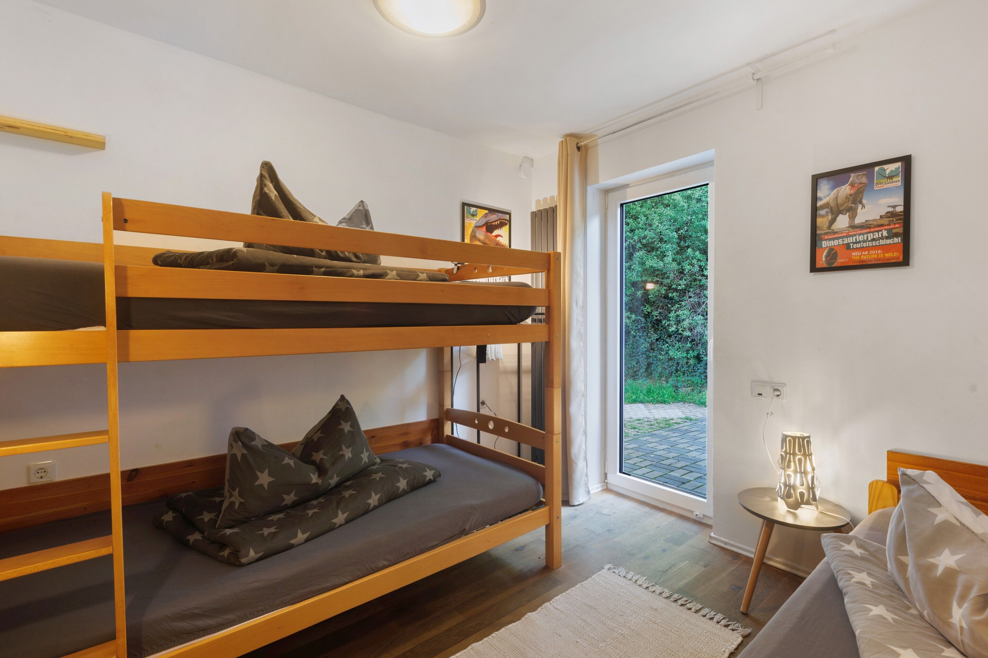 A bunk bed room with a window, a small table with a lamp, and a poster on the wall.