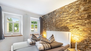 A bedroom with a stone wall, a bed with patterned pillows, a bedside table with a lamp, and a window with curtains. - Holiday Home 'Enztal' with Shared Terrace (Zweifelscheid)