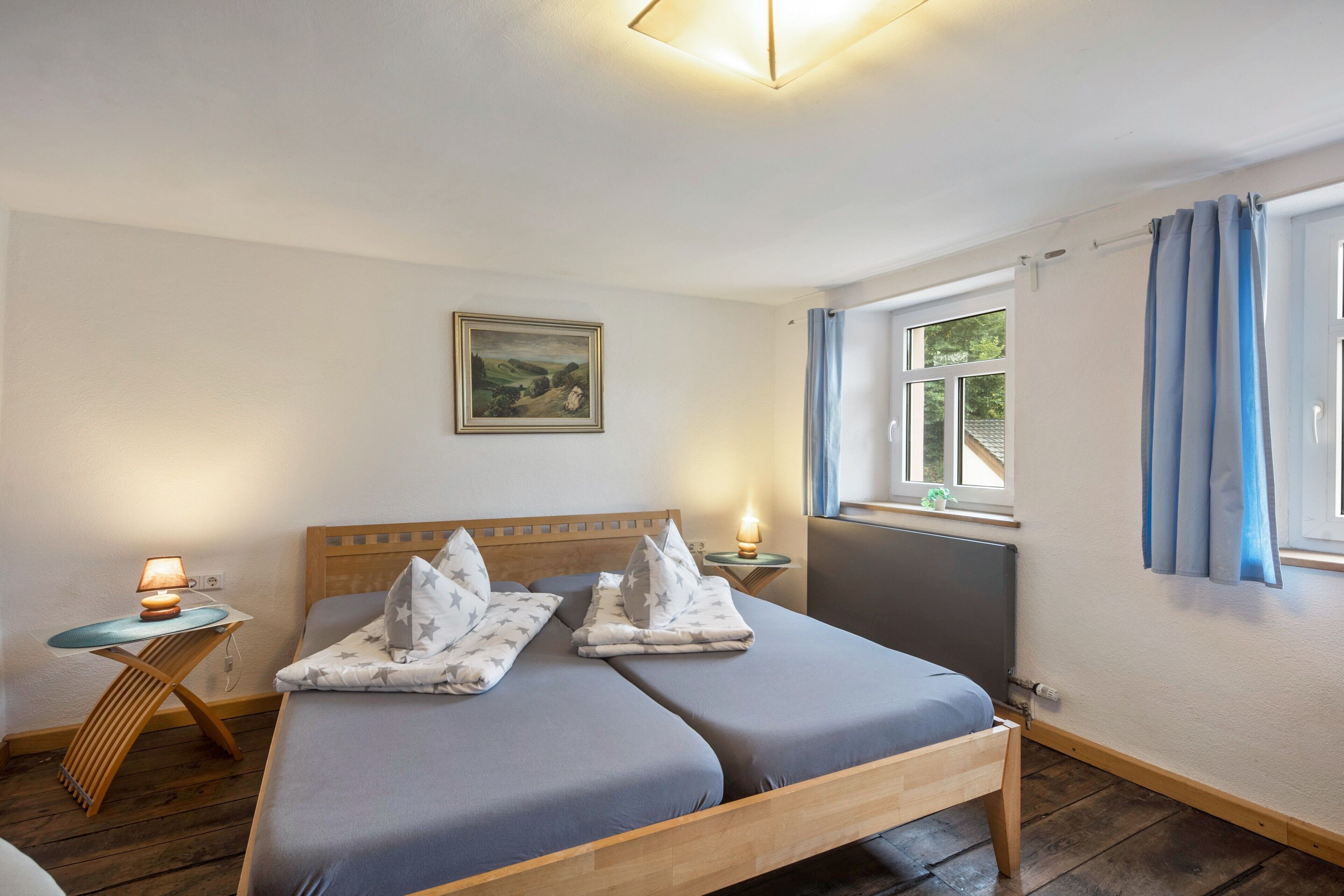 Ein Doppelbett mit Holzrahmen, ein Nachttisch mit Lampe, ein Fenster mit Vorhängen und ein Bild an der Wand.