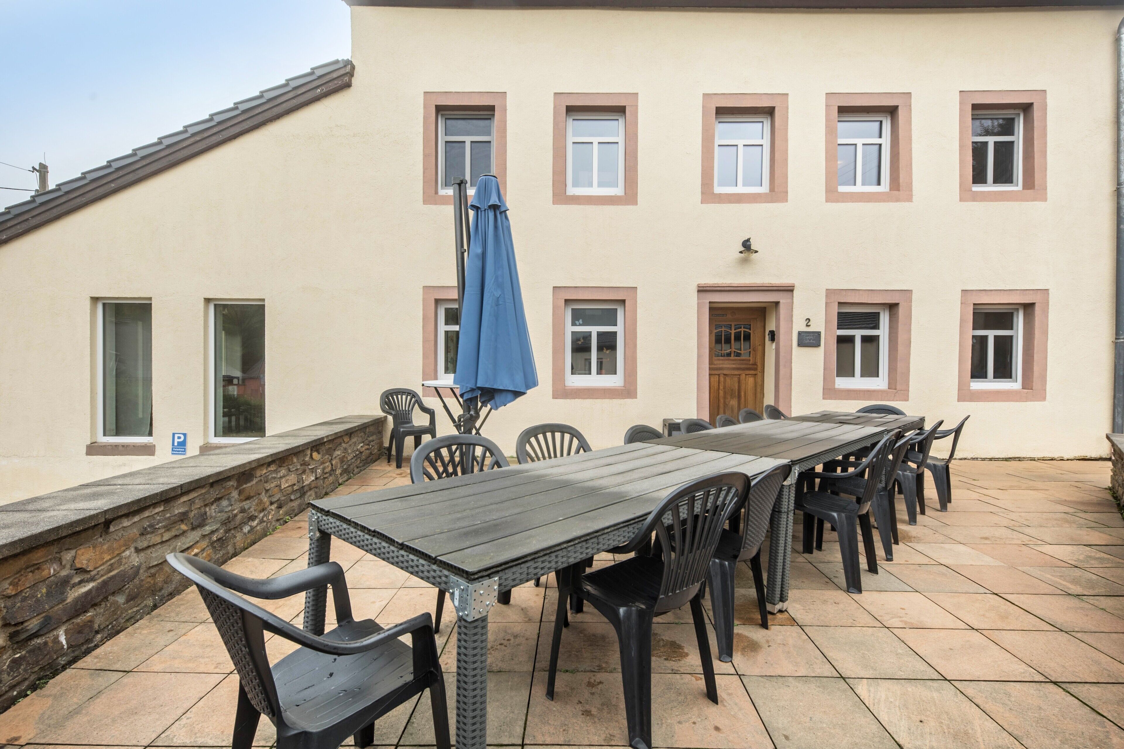 An outdoor patio with a long table and chairs, a stone wall, and a building with multiple windows and a door.