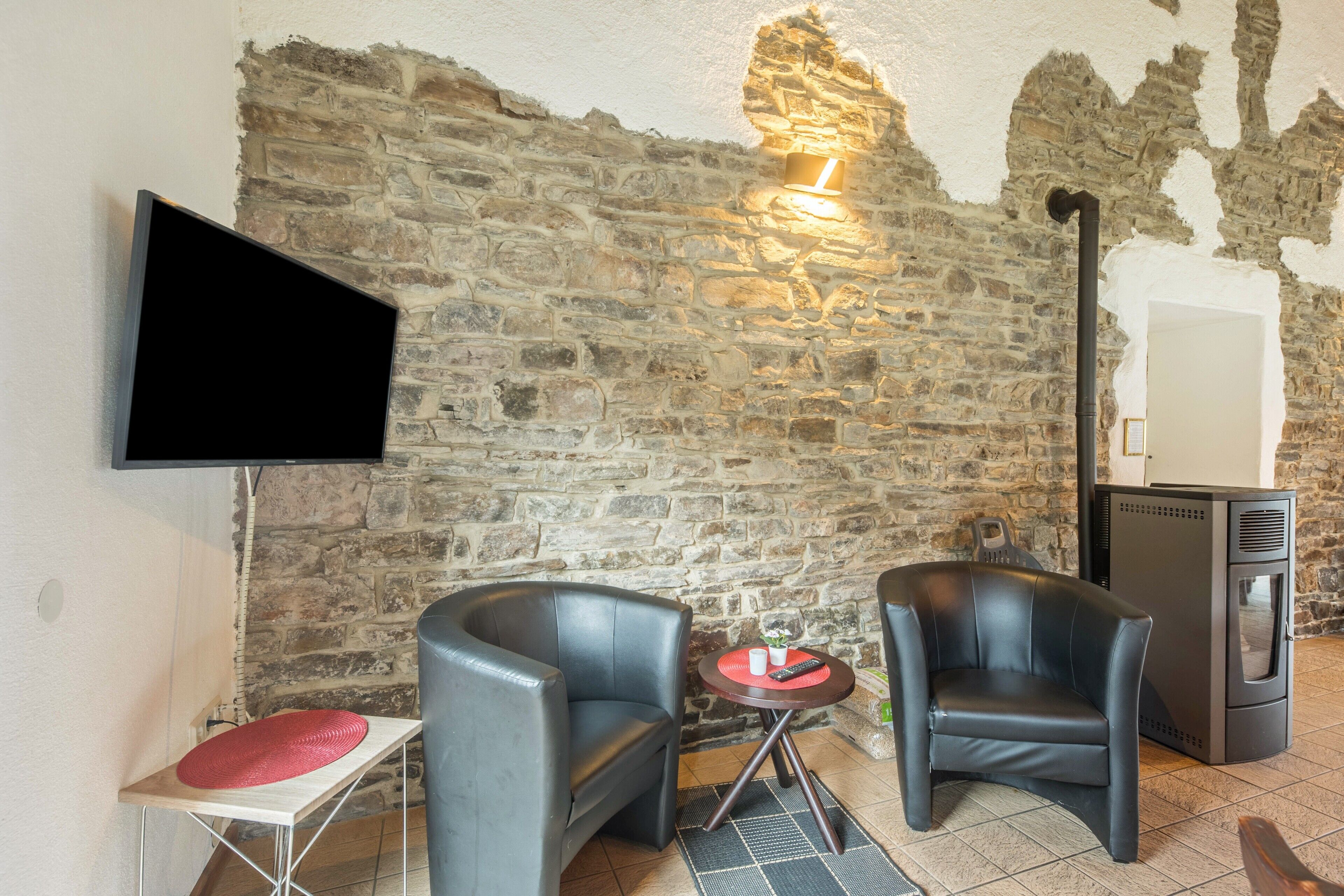 Une pièce avec un mur en pierre, un téléviseur à écran plat fixé au mur, deux fauteuils en cuir, une petite table ronde avec un coussin rouge et un poêle à bois.