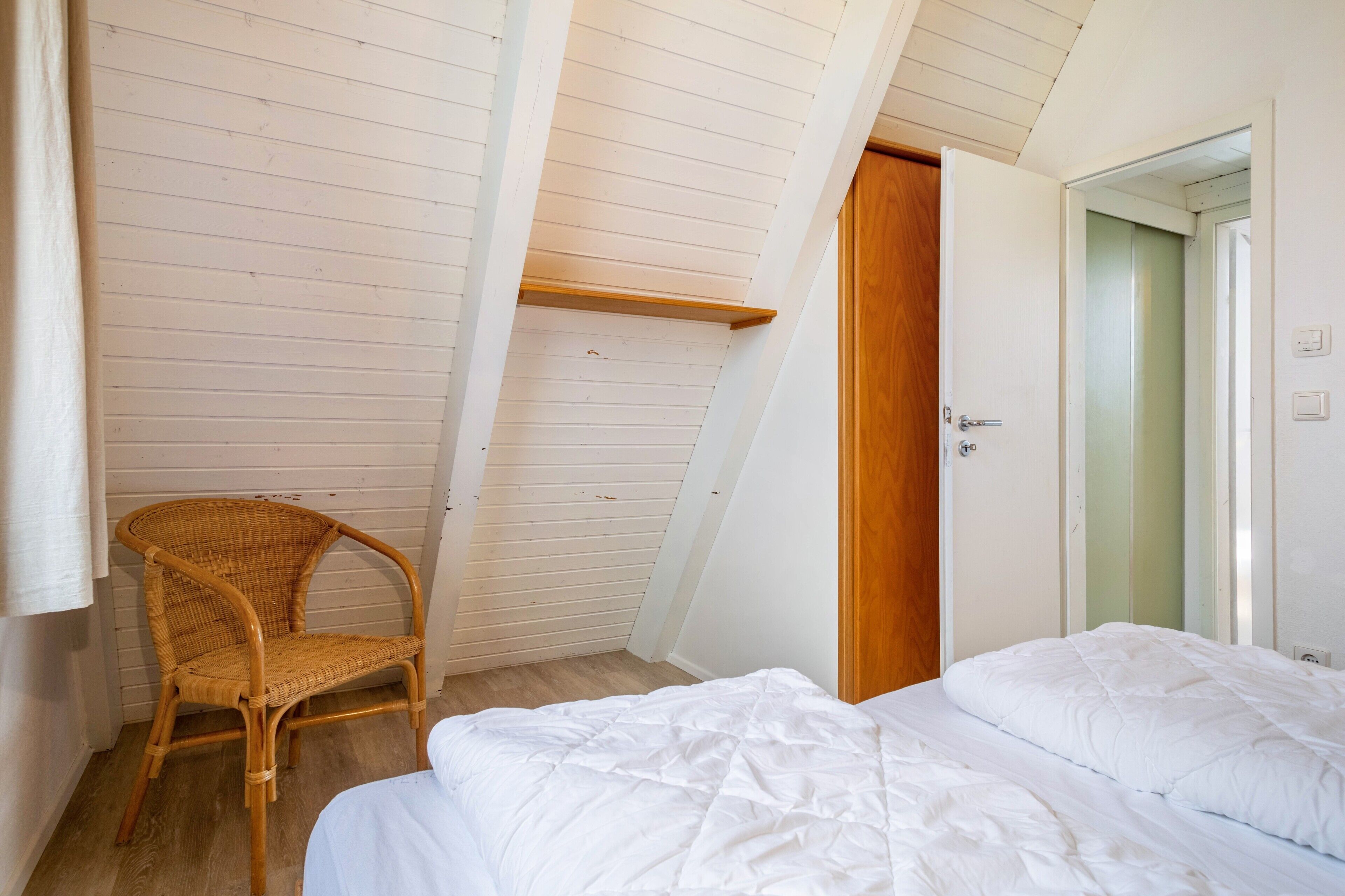 A bedroom with a bed, a wooden chair, a slanted ceiling, and a door.