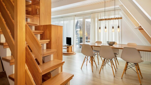 A modern interior with a wooden staircase, a dining area with chairs and a table, and a large window with curtains.