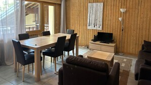 Een moderne woonkamer met houten vloer, een eetgedeelte met een houten tafel en stoelen, een flatscreen televisie op een houten kast en een groot raam met gordijnen.