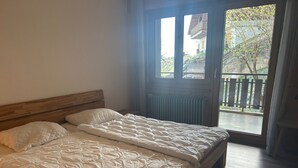 Een slaapkamer met een houten bed, een raam met uitzicht op een balkon en bomen, en een radiator onder het raam.
