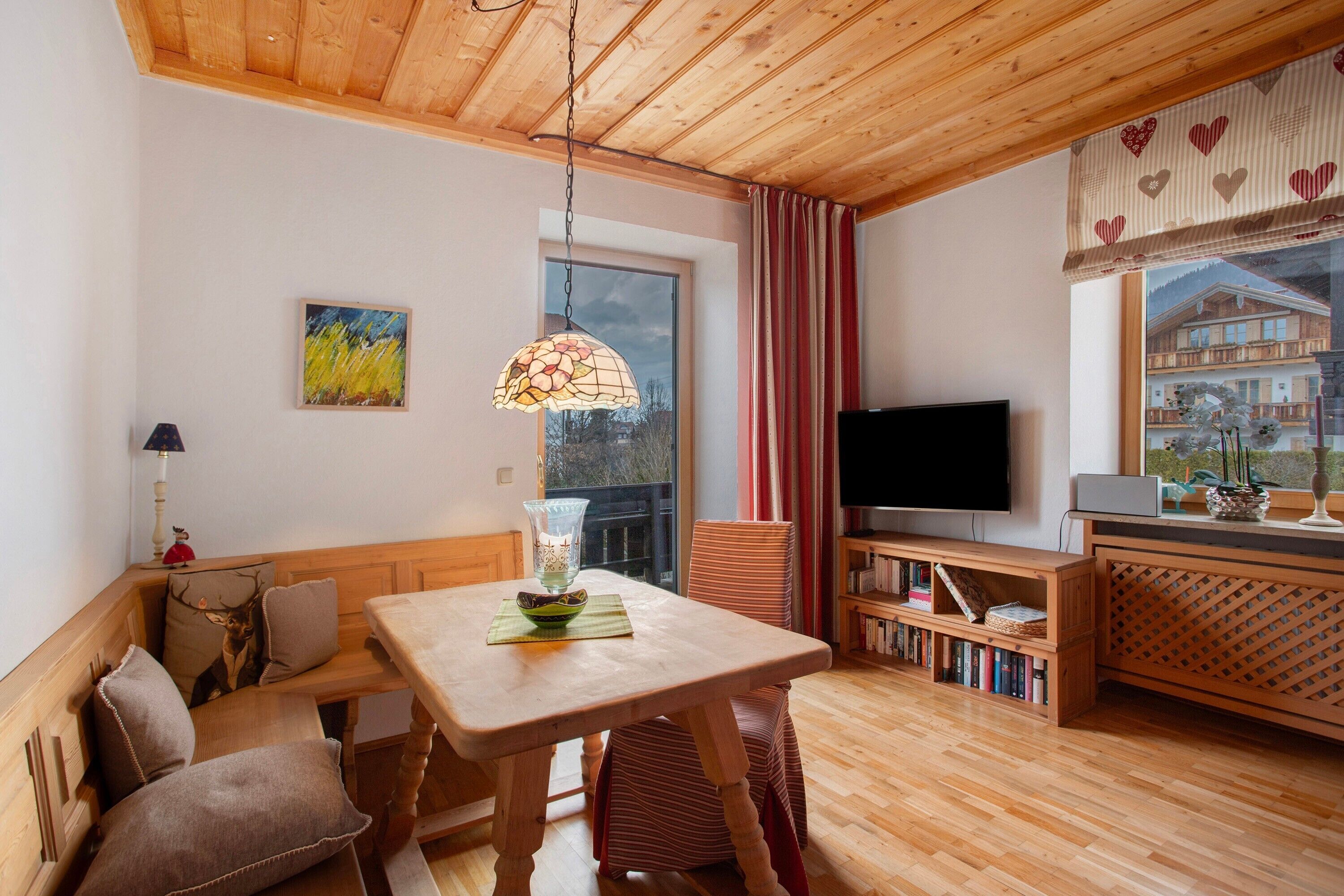 Ein gemütliches Zimmer mit Holzboden, einem Esstisch, einer Bank, einem Fernseher und einem Bücherregal.