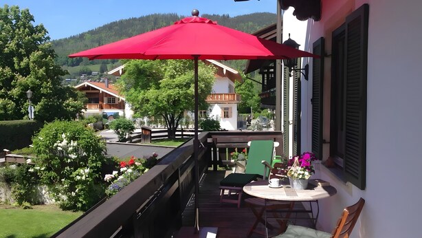 Un balcón con una sombrilla roja, una mesa con sillas y vistas a un pueblo con casas y árboles.