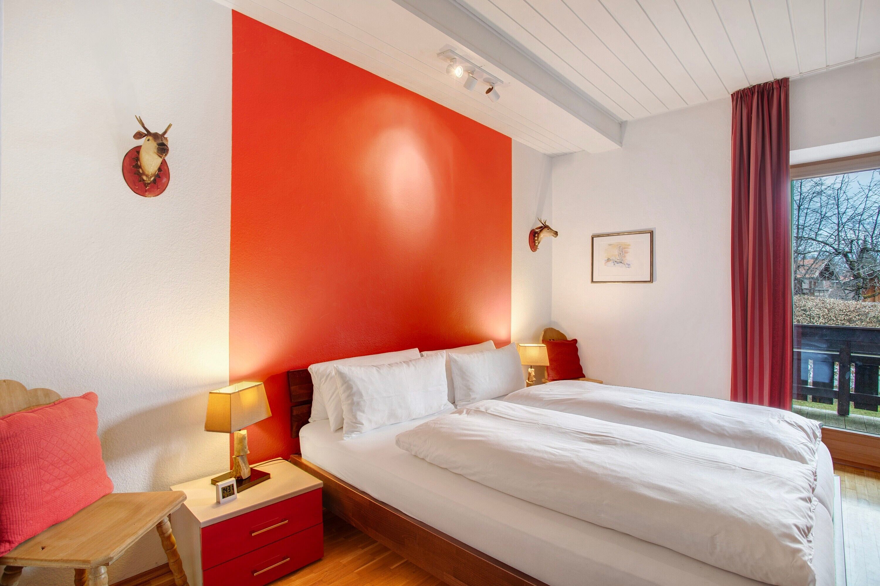 Ein Schlafzimmer mit einem großen Bett, zwei Nachttischen, einem Schreibtisch und einer roten Wand, sowie einem Fenster mit Blick.