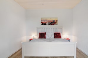 Una cama blanca con almohadas rojas, un cabecero blanco y un cuadro de un muelle en la pared.