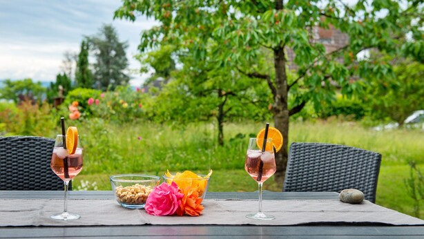 Two glasses of pink beverage with orange slices on a table with a wicker chair, a bowl of nuts, and a flower.