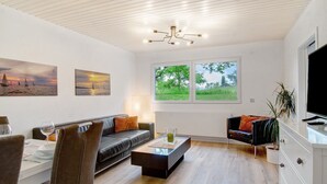 A modern living room with a sofa, dining table, and chairs, a coffee table, a TV, and a large window with a view of greenery.