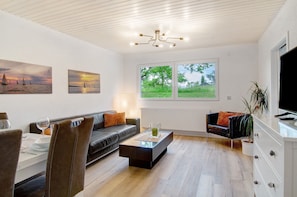 Un salon moderne comprenant un canapé, une table à manger et des chaises, une table basse, un téléviseur et une grande fenêtre donnant sur un paysage verdoyant.