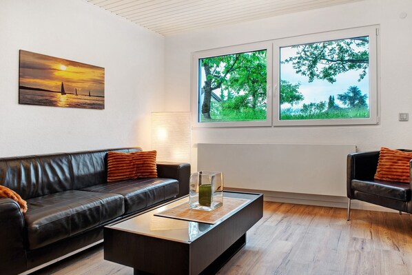A modern living room with a black leather sofa, a glass coffee table, and a large window with a view of trees. - Holiday Apartment 'With Garden View' (Uhldingen-Mühlhofen)