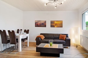 Una sala de estar moderna con comedor, un sofá de cuero negro, una mesa de centro de vidrio y dos cuadros de paisajes al atardecer en la pared.