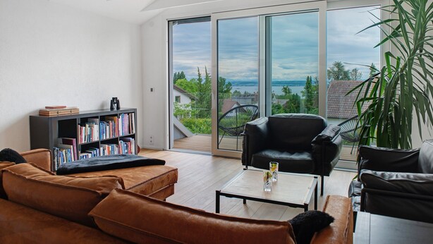A modern living room with a large sliding glass door leading to a balcony, a leather sofa, a black armchair, a coffee table, and a bookshelf.