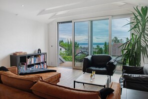 Un salon moderne doté d’une grande porte-fenêtre coulissante donnant sur un balcon, d’un canapé en cuir, d’un fauteuil noir, d’une table basse et d’une bibliothèque.