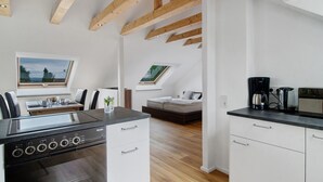 A modern kitchen with a built-in stove, wooden beams, a dining area with a table and chairs, and a bedroom with a bed and windows.