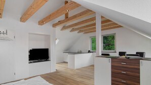 A modern loft bedroom with exposed wooden beams, a large window, and a kitchenette.