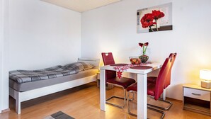 Una habitación pequeña con una cama, una mesa de comedor con sillas, una mesita con una lámpara y un cuadro en la pared.