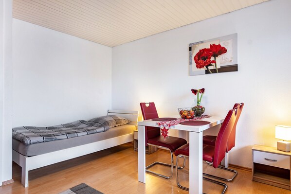 A small room with a bed, a dining table with chairs, a small table with a lamp, and a painting on the wall.