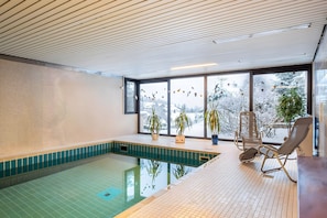 Une piscine intérieure avec un sol carrelé, de grandes fenêtres et des plantes en pot.