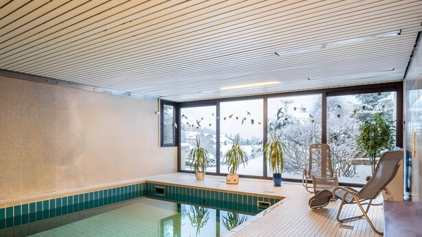Una piscina interior con suelo de baldosas, ventanas amplias y plantas en macetas.
