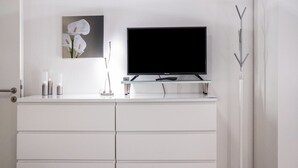 Un baño moderno con un tocador blanco, un televisor de pantalla plana, un estante de vidrio, un cuadro de flores blancas y una decoración minimalista.
