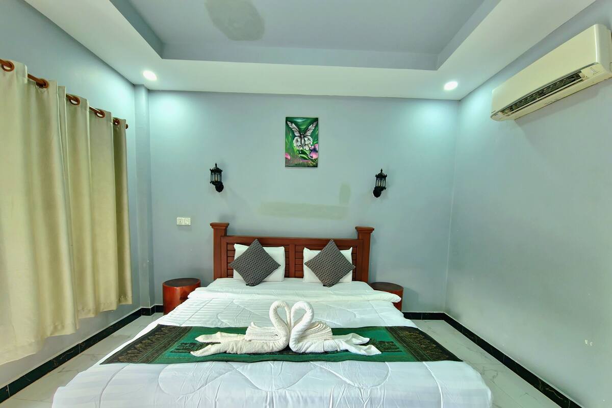 A neatly made bed with a swan-shaped pillow and a green mat, a wooden headboard, two wall-mounted lamps, and an air conditioning unit on the wall.