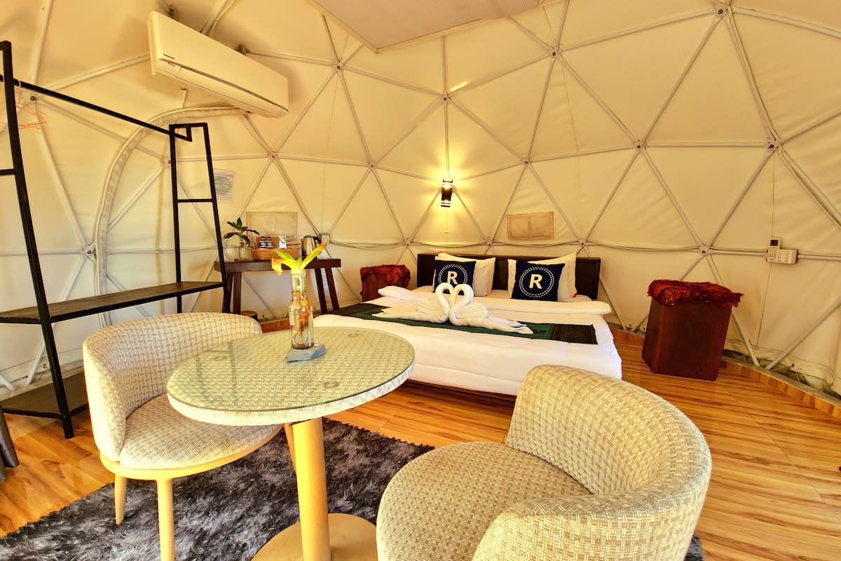 A tented accommodation with a bed, two chairs, a table, and a small plant.