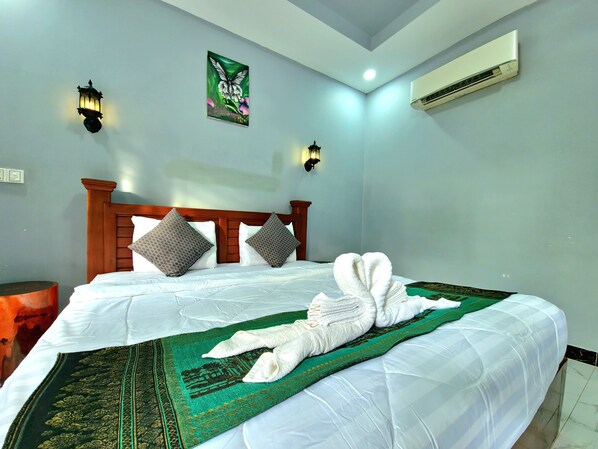 A hotel room with a neatly made bed, a green and gold patterned bedspread, two pillows, a wall-mounted air conditioner, and a painting of a butterfly on the wall. - Phnom Tbeng Resort (Tbaeng Meanchey)